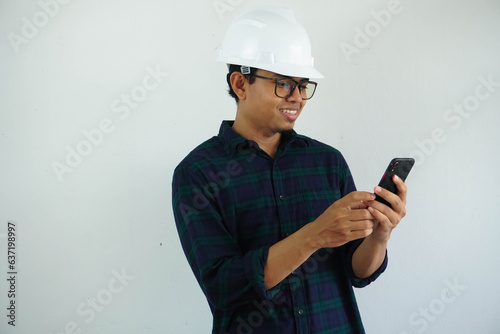 smiling young Asian male engineer wearing white hard hat holding mobile phone for construction work isolated on white background.