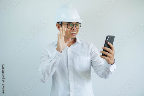 smiling young asian man architect wearing white helmet hard hat waving hand with happy face when doing video call with someone isolated on white background. photo