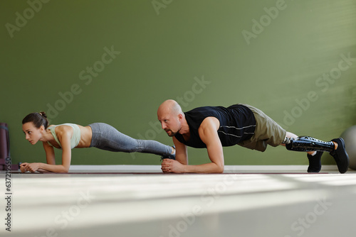 Portrait of sportive young couple working out indoors with man with prosthetic leg doing plank exercise photo