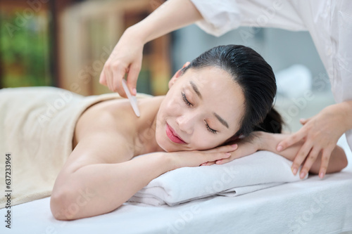 아시아 한국의 젊은 여자 모델이 스파 또는 피부관리전문점 또는 마사지전문점에서 침대에 엎드려 베이지색 대형 타올을 덮고 하얀색 셔츠를 입은 마사지전문가 또는 피부전문관리사가 목,어깨,얼굴,등 상반신에 오일 또는 기능성 화장품을 바르고있다.