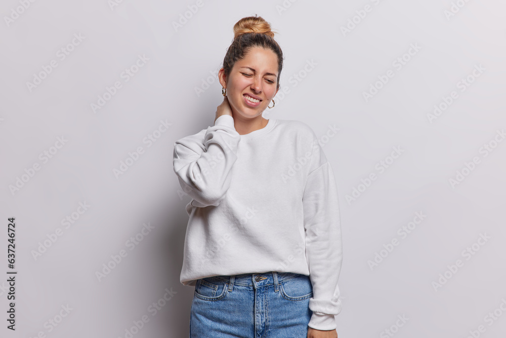 Discontent young woman touching neck suffers from muscle tension clenches teeth needs massage wears casual sweatshirt and jeans poses against white background works lot in front of computer. Discontent young woman touching neck suffers from muscle tension clenches teeth needs massage wears casual sweatshirt and jeans poses against white background works lot in front of computer.