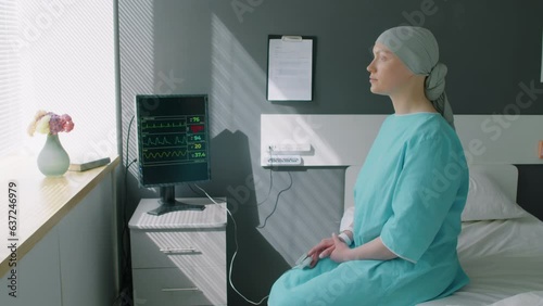 Young female depressive patient sitting on bed in ward after chemo and looking at window in hospital photo