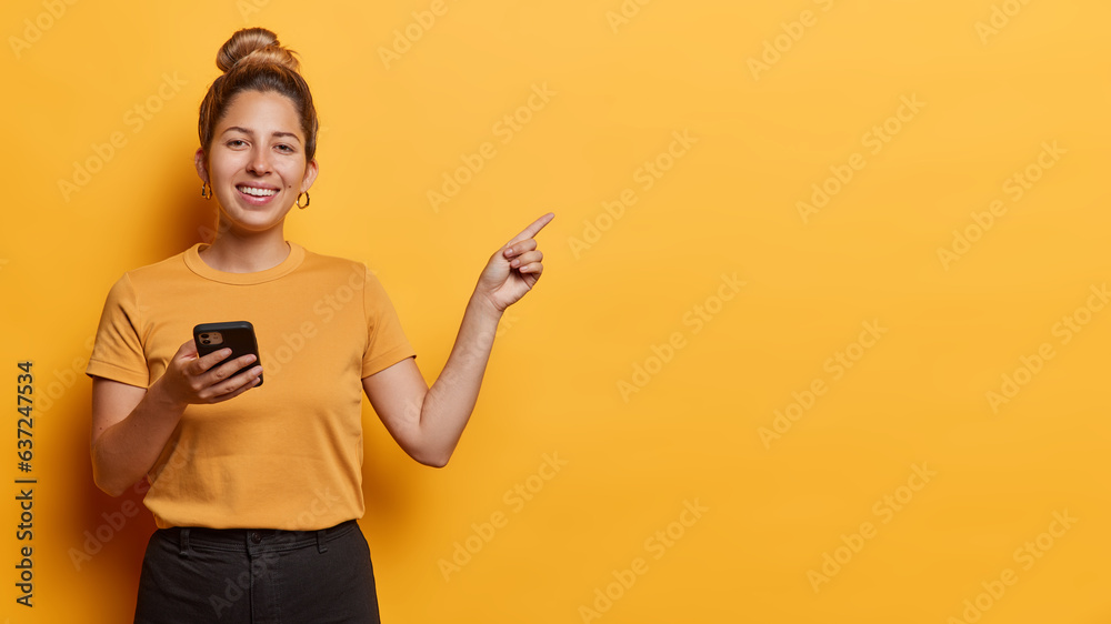 Horizontal shot of pleasant looking cheerful woman holds mobile phone for chatting online points index finger on empty space isolated over yellow background. People and advertisement concept Horizontal shot of pleasant looking cheerful woman holds mobile phone for chatting online points index finger on empty space isolated over yellow background. People and advertisement concept