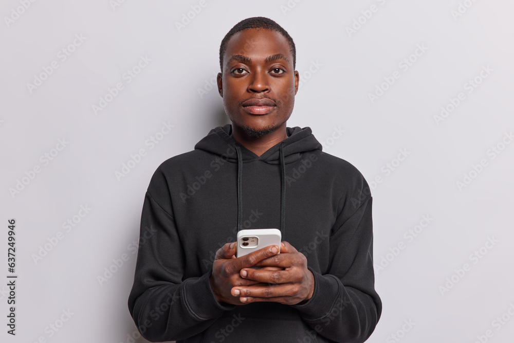 People positive emotions concept. Studio shot of young happy African american man holding smartphone wearing black hoodie standing in centre isolated on white background looking straight at camera