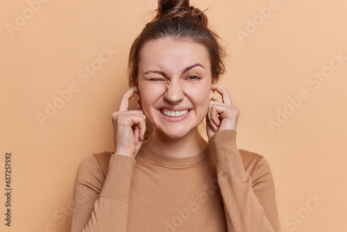 Negative emotions concept. Studio shot of young stressed strained European girl standing isolated in centre on beige background plugging ears not wanting to hear unpleasnat loud sounds or noise