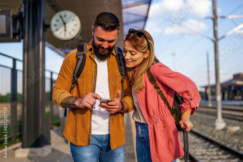 Happy couple is standing at railway station and waiting for arrival of their train. They are looking at map on smartphone.