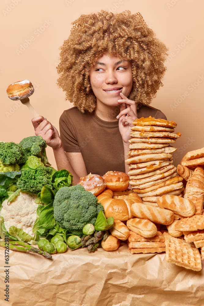Thoughtful woman with curly hair poses near table full of helthy and unhealthy food holds fork with doughnut looks aside contemplates about something wears casual t shirt poses over brown background Thoughtful woman with curly hair poses near table full of helthy and unhealthy food holds fork with doughnut looks aside contemplates about something wears casual t shirt poses over brown background
