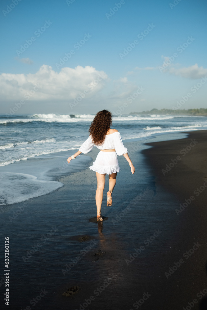 A young, beautiful girl, with curly hair, in white clothes, walks along the beach and enjoys life, rear view.