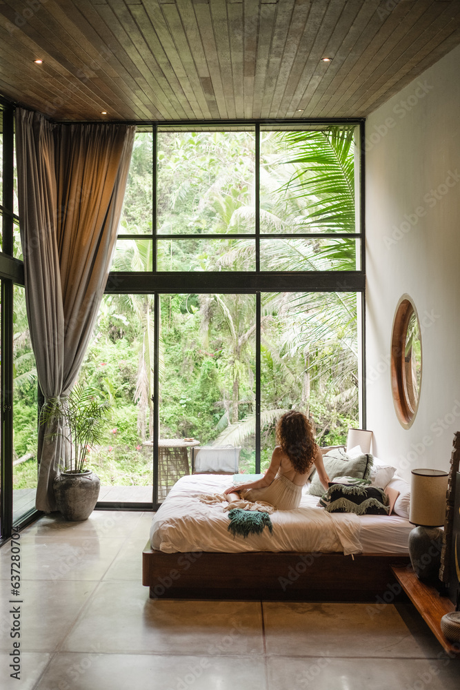 A girl of European appearance, with curly hair, greets the morning in a luxurious villa on the bed, admiring the nature outside the large window. - obrazy, fototapety, plakaty 