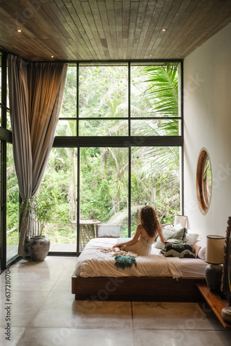 A girl of European appearance, with curly hair, greets the morning in a luxurious villa on the bed, admiring the nature outside the large window. © Evgenii