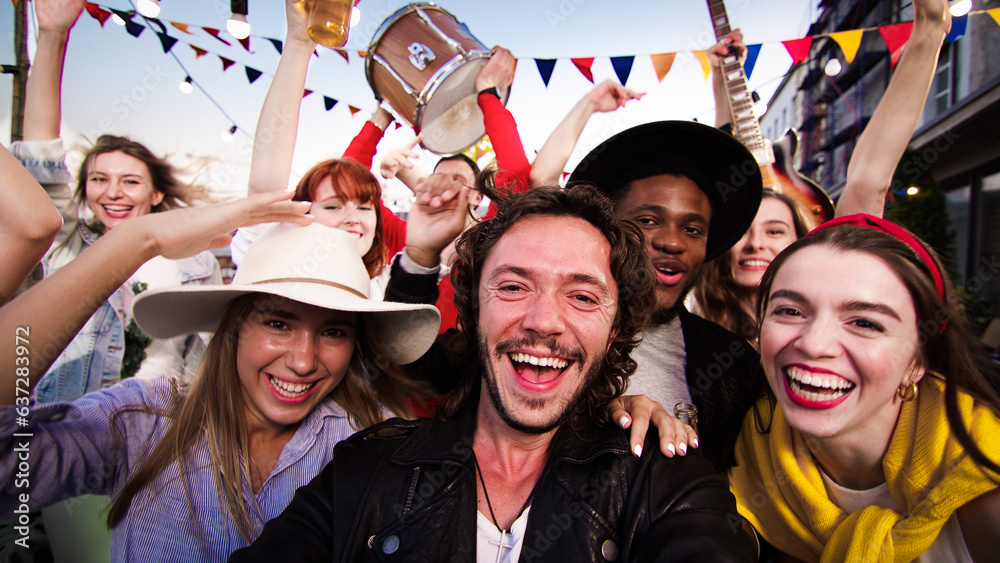 Fototapeta premium Young caucasian man making video with his friends on a party using smartphone. Rooftop party friends having fun, singing and dancing together.