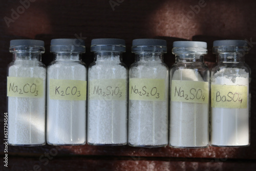 White inorganic reagents in glass tubes: sodium and potassium carbonates, sodium silicate, sodium thiosulfate, sodium and barium sulfates are in a wooden table.  photo