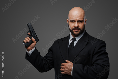 Bald security man in a suit holds a gun, ready to protect, against a gray studio backdrop