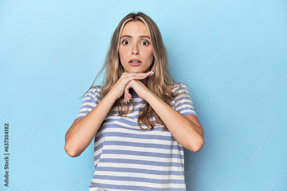 Blonde young caucasian woman in blue studio praying for luck, amazed and opening mouth looking to front. Blonde young caucasian woman in blue studio praying for luck, amazed and opening mouth looking to front.