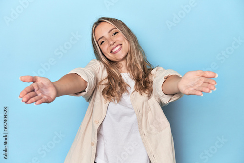 Blonde young caucasian woman in blue studio showing a welcome expression.