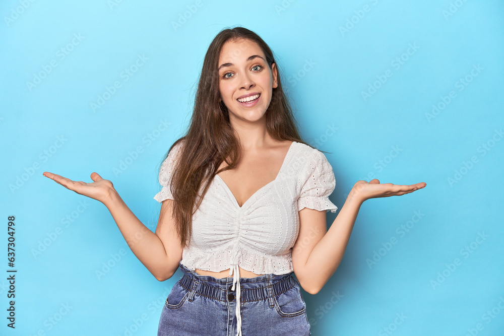 Stylish young woman in white blouse on a blue studio backdrop makes scale with arms, feels happy and confident. Stylish young woman in white blouse on a blue studio backdrop makes scale with arms, feels happy and confident.