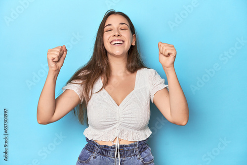 Stylish young woman in white blouse on a blue studio backdrop celebrating a victory, passion and enthusiasm, happy expression.