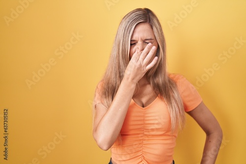 Young woman standing over yellow background smelling something stinky and disgusting, intolerable smell, holding breath with fingers on nose. bad smell