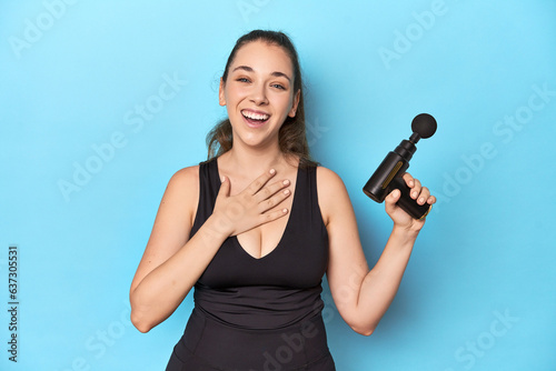 Active woman with an electric massager in a blue studio laughs out loudly keeping hand on chest.