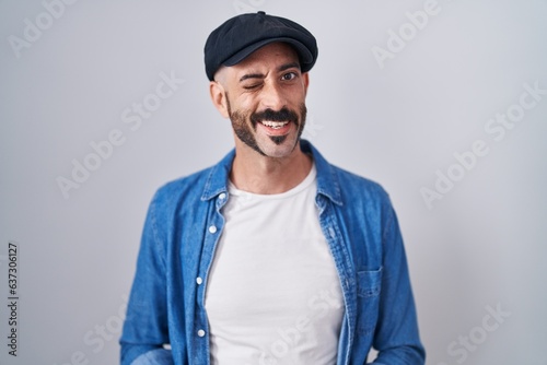 Hispanic man with beard standing over isolated background winking looking at the camera with sexy expression, cheerful and happy face.