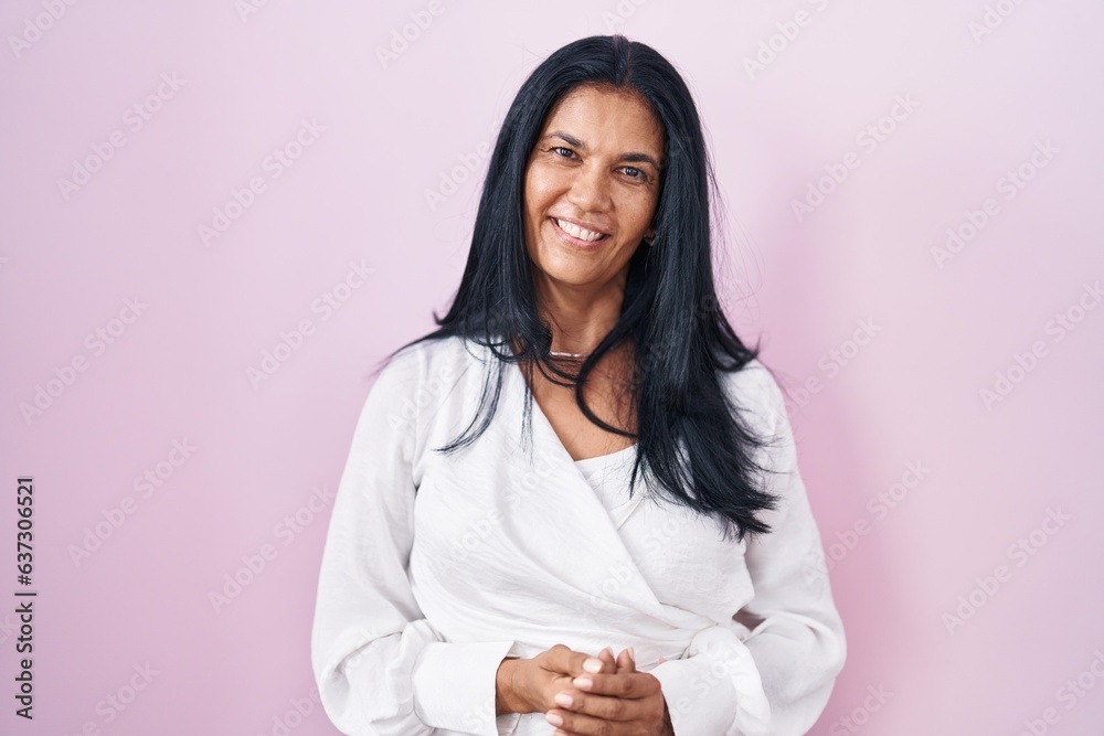 Mature hispanic woman standing over pink background with hands together and crossed fingers smiling relaxed and cheerful. success and optimistic Mature hispanic woman standing over pink background with hands together and crossed fingers smiling relaxed and cheerful. success and optimistic