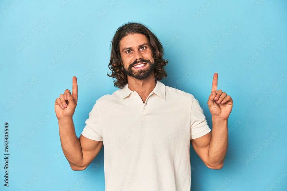 Long-haired man in a white polo, blue studio indicates with both fore fingers up showing a blank space. Long-haired man in a white polo, blue studio indicates with both fore fingers up showing a blank space.