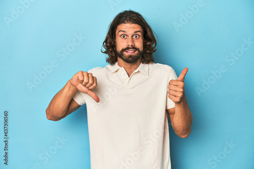 Long-haired man in a white polo  blue studio showing thumbs up and thumbs down  difficult choose concept