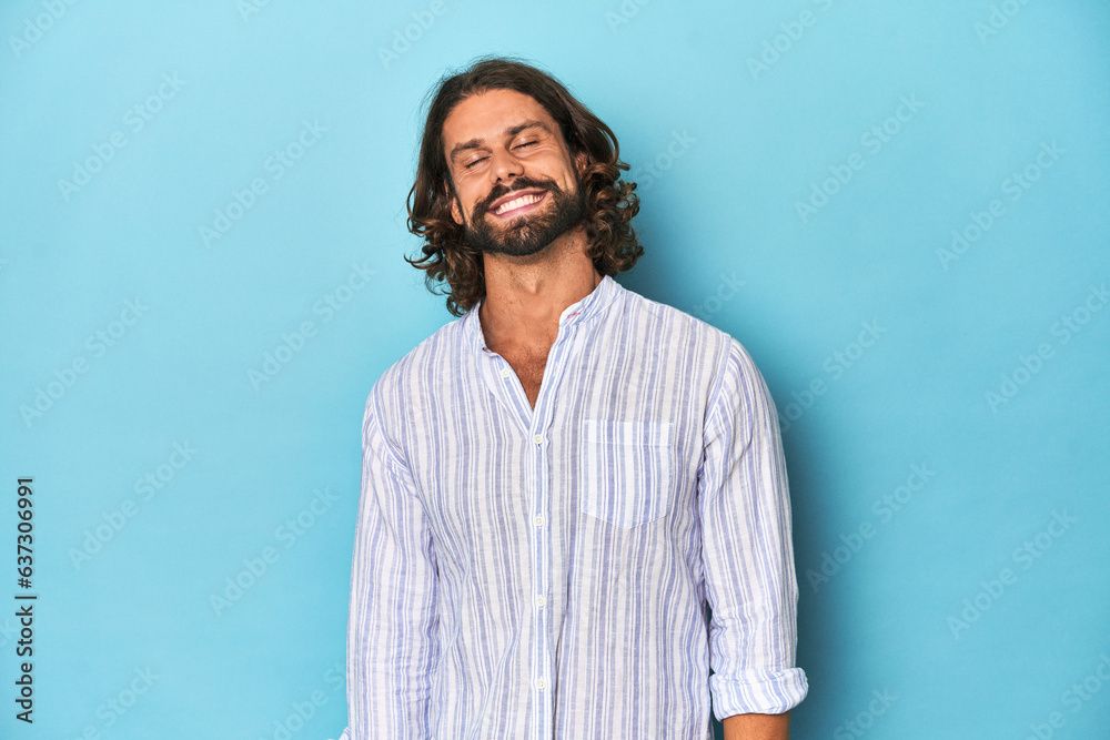 Fototapeta premium Man with beard in blue striped shirt, blue studio laughs and closes eyes, feels relaxed and happy.