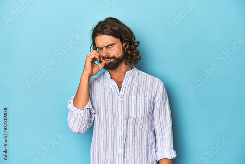 Man with beard in blue striped shirt, blue studio with fingers on lips keeping a secret.