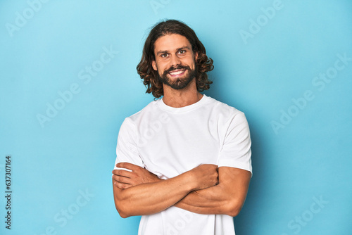 Bearded man in a white shirt, blue backdrop who feels confident, crossing arms with determination.