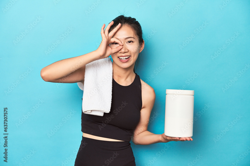 Young Asian sportswoman with protein bottle, gym setup, excited keeping ok gesture on eye. Young Asian sportswoman with protein bottle, gym setup, excited keeping ok gesture on eye.
