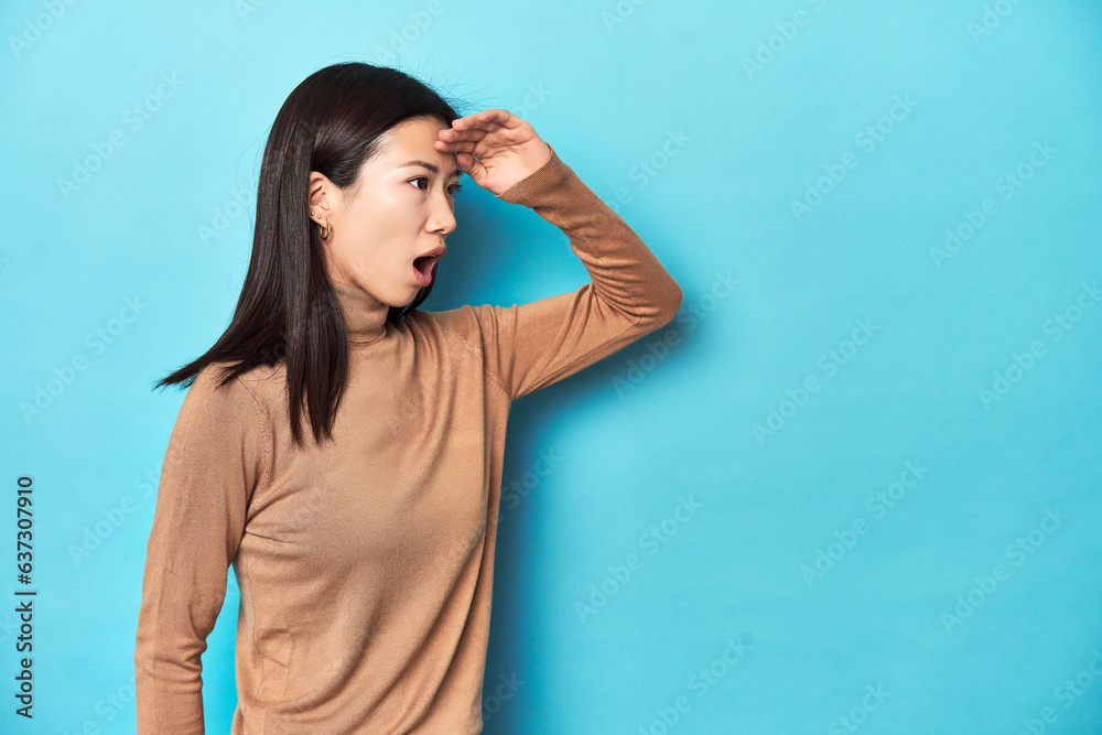 Young Asian woman in brown turtleneck, looking far away keeping hand on forehead.