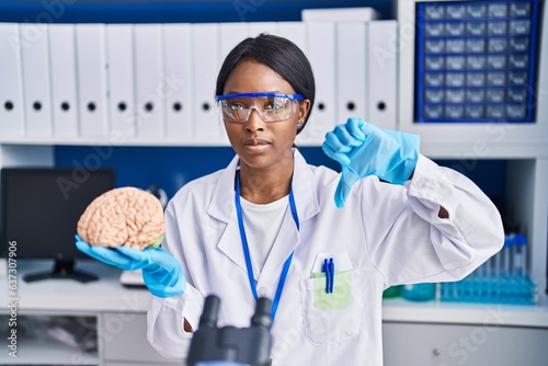 African young woman working at scientist laboratory holding brain with angry face  negative sign showing dislike with thumbs down  rejection concept