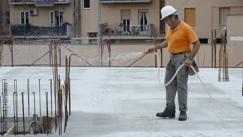 Construction worker watering fresh concrete slab using a hose.