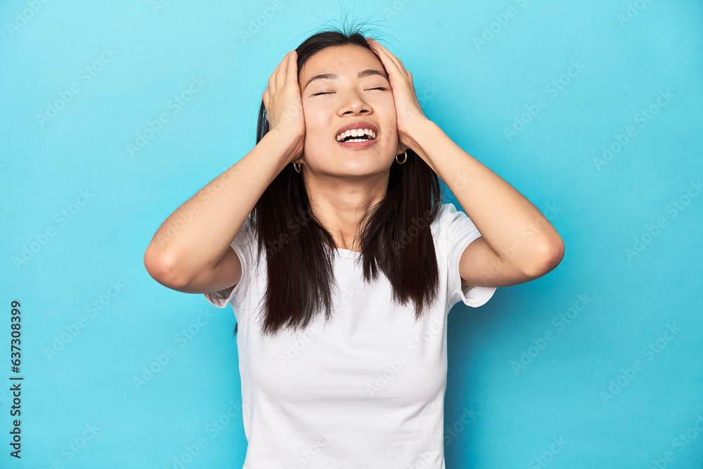 Young Asian woman in white t-shirt, studio shot, laughs joyfully keeping hands on head. Happiness concept. Young Asian woman in white t-shirt, studio shot, laughs joyfully keeping hands on head. Happiness concept.