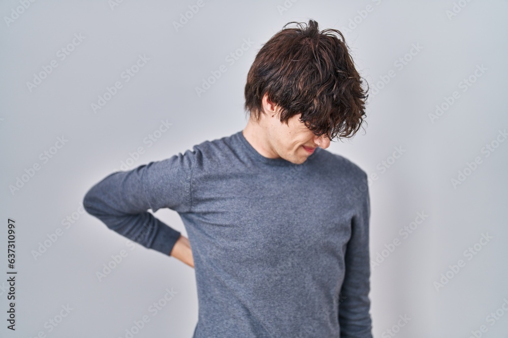 Young man standing over isolated background suffering of backache, touching back with hand, muscular pain Young man standing over isolated background suffering of backache, touching back with hand, muscular pain