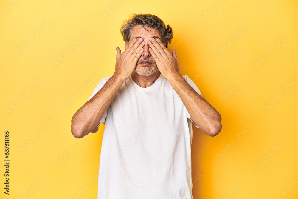 Middle-aged man posing on a yellow backdrop afraid covering eyes with hands. Middle-aged man posing on a yellow backdrop afraid covering eyes with hands.