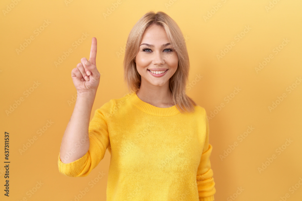 Young caucasian woman wearing yellow sweater showing and pointing up with finger number one while smiling confident and happy. Young caucasian woman wearing yellow sweater showing and pointing up with finger number one while smiling confident and happy.