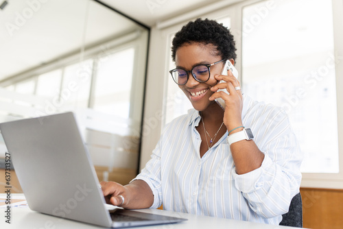 Confident young entrepreneur using laptop, talking on mobile phone in contemporary co-working space. Stylish african-american woman with short haircut sitting at desk, has pleasant phone conversation