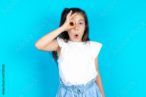 Caucasian kid girl wearing  white t-shirt over blue background doing ok gesture shocked with surprised face, eye looking through fingers. Unbelieving expression. © Roquillo