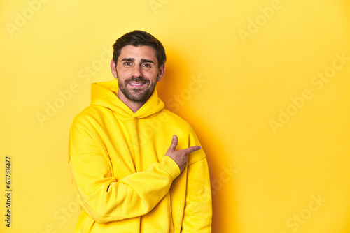 Man in yellow hoodie, monochrome studio backdrop smiling and pointing aside, showing something at blank space.