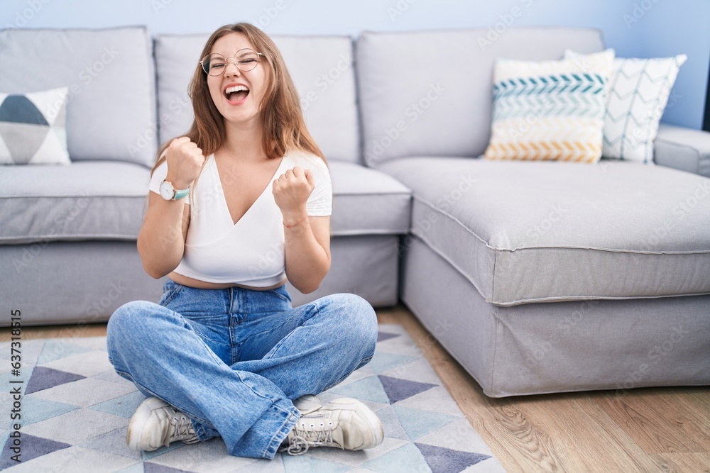 Young caucasian woman sitting on the floor at the living room very happy and excited doing winner gesture with arms raised, smiling and screaming for success. celebration concept. Young caucasian woman sitting on the floor at the living room very happy and excited doing winner gesture with arms raised, smiling and screaming for success. celebration concept.