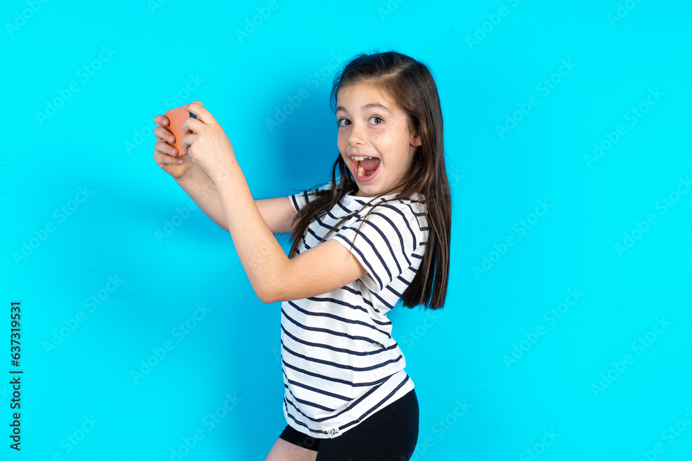 Nice addicted cheerful Caucasian kid girl wearing floral dress over blue background using gadget playing network game Nice addicted cheerful Caucasian kid girl wearing floral dress over blue background using gadget playing network game