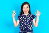 Optimistic Caucasian kid girl wearing floral dress over blue background  raises palms from joy, happy to receive awesome present from someone, shouts loudly,