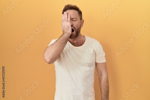 Middle age man with beard standing over yellow background yawning tired covering half face, eye and mouth with hand. face hurts in pain.