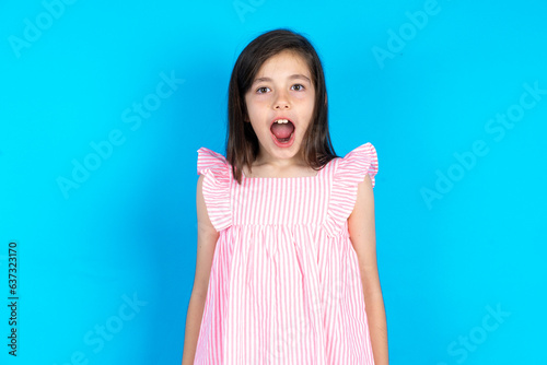 Oh my God. Surprised caucaisna kid girl wearing pink dress over blue background stares at camera with shocked expression exclaims with unexpectedness, photo