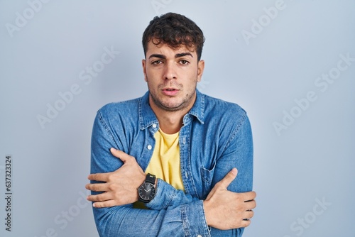 Photos Young hispanic man standing over blue background shaking and freezing for winter
