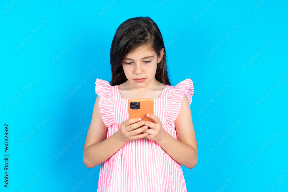 Focused caucaisna kid girl wearing pink dress over blue background use smartphone reading social media news, or important e-mail Focused caucaisna kid girl wearing pink dress over blue background use smartphone reading social media news, or important e-mail