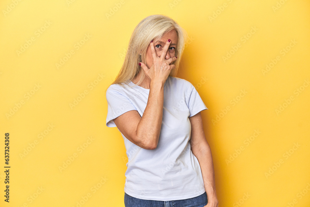 Middle-aged Caucasian woman in white t-shirt, yellow studio blink at the camera through fingers, embarrassed covering face. Middle-aged Caucasian woman in white t-shirt, yellow studio blink at the camera through fingers, embarrassed covering face.