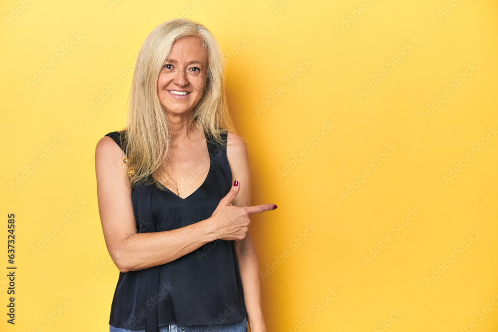 Elegant middle-aged woman in black top, yellow studio smiling and pointing aside, showing something at blank space. Elegant middle-aged woman in black top, yellow studio smiling and pointing aside, showing something at blank space.